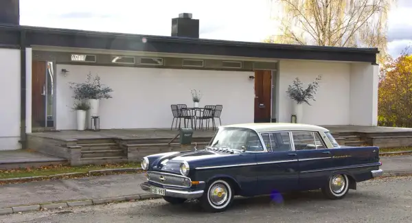 En mörkblå Opel Kapitän från 1960 står parkerad framför ett modernt hus med träaltan.