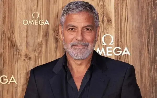 Un homme avec des cheveux gris courts et une barbe, portant un costume sombre, pose devant un mur en bois avec le logo Omega.