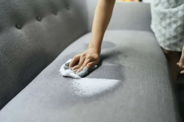 A hand with a soapy cloth gently cleans a grey couch cushion, showing how to wash couch cushion covers for a fresh look.