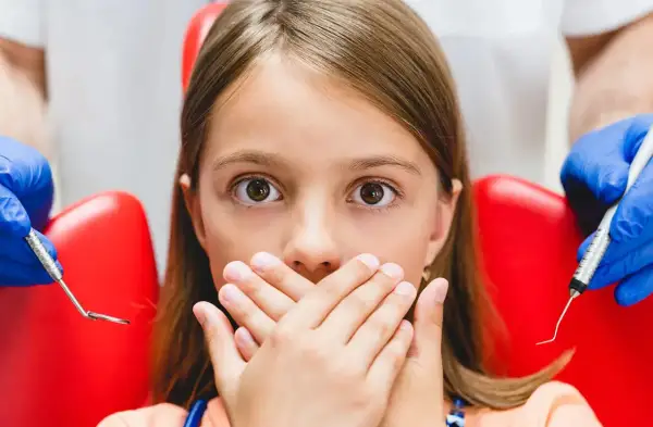 Przerażona dziewczynka zakrywa usta dłońmi, gdy dentysta przygotowuje się do podania znieczulenia. Wokół niej widać czerwony fotel dentystyczny i narzędzia.