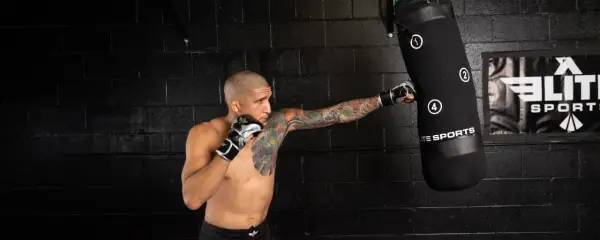 A tattooed man in boxing gloves performs a punching bag workout, his fist connecting with the bag.