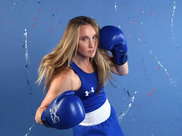 A determined female boxer in blue gear, showcasing the spirit of amateur boxing against a vibrant, paint-splattered background.