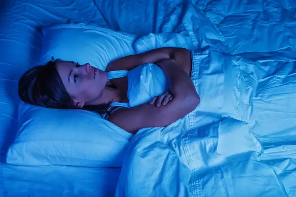 Frau liegt wach im Bett, Arme verschränkt, die Augen weit aufgerissen. Deutliche Anzeichen von Schlafmangel und dessen Folgen.