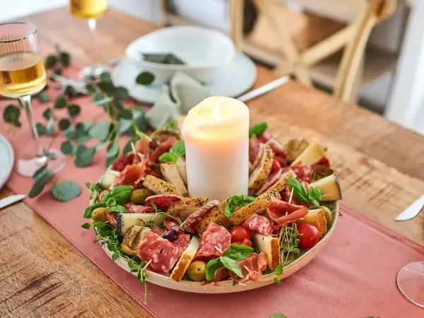 Antipasti selber machen: Eine festliche Platte mit Wurst, Käse, Brot, Oliven und Tomaten, umgeben von grünen Zweigen, mit einer Kerze in der Mitte.