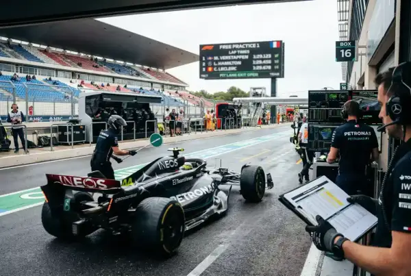 Coche de F1 en boxes durante entrenamientos. Un miembro del equipo guía al piloto mientras otros revisan tiempos en pantalla.