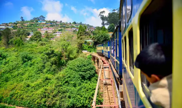 Ett blått och gult tåg i Indien färdas längs en bergssluttning täckt av frodig grönska och färgglada hus.