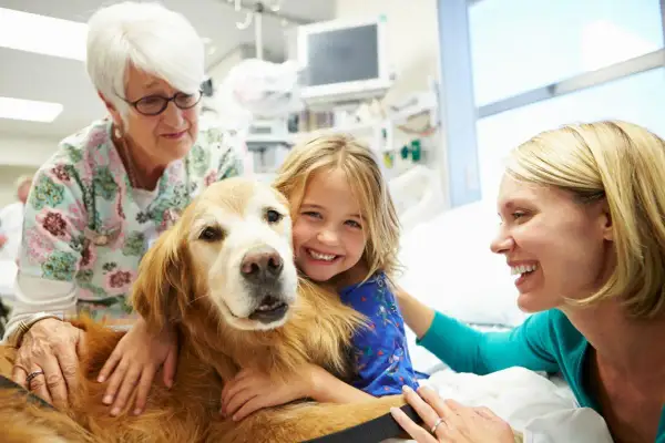 Niña sonriente abraza a un perro de terapia mientras una enfermera y su madre observan.