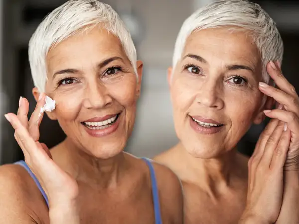 Zwei Frauen mit kurzen weißen Haaren lächeln. Eine trägt Creme auf ihre Wange auf, um trockene Haut im Alter zu bekämpfen.