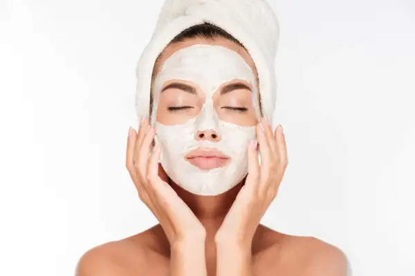 Une femme avec une serviette sur la tête applique un masque blanc sur son visage, espérant atténuer les marques de masque cicatrice acné.