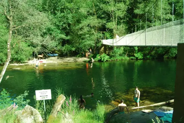 Puente colgante sobre río verde con gente bañándose y disfrutando del paisaje.