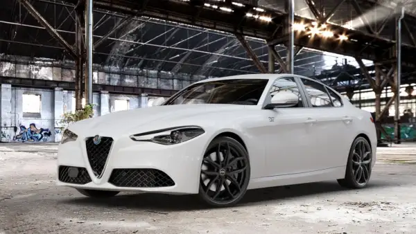 Biała Alfa Romeo Giulia Veloce z czarnymi felgami stoi w opuszczonej hali przemysłowej, gdzie światło przebija się przez dach.