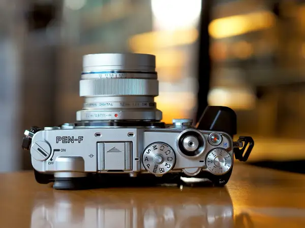 Silver Olympus PEN-F camera with a silver Olympus Digital 12mm 1:2.0 lens attached. The camera body features various dials and buttons, ready for capturing stunning images with its Olympus PEN F lenses.
