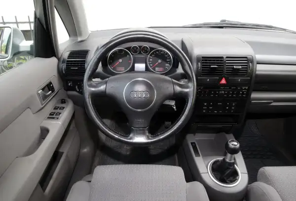 Wnętrze Audi A2 z kierownicą, deską rozdzielczą i dźwignią zmiany biegów. Widoczne wskaźniki, radio i przyciski.