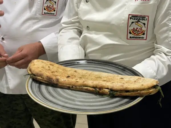 Una paposcia pugliese, ripiena e dorata, servita su un piatto grigio. Indossano giacche con stemmi dell'Associazione Pizzaioli Garganici.