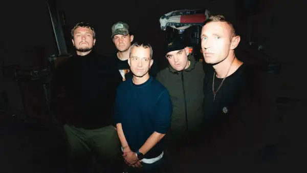 Five men stand in a dimly lit room, ready to perform. One holds a high vis setlist, hinting at the night's energetic show.