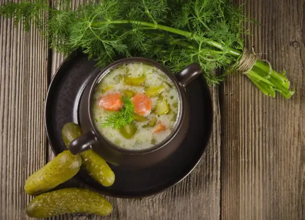 Aromaticzna zupa ogórkowa na żeberkach, podana w brązowej miseczce, ozdobiona koperkiem. Obok leżą chrupiące ogórki kiszone i pęczek świeżego koperku.
