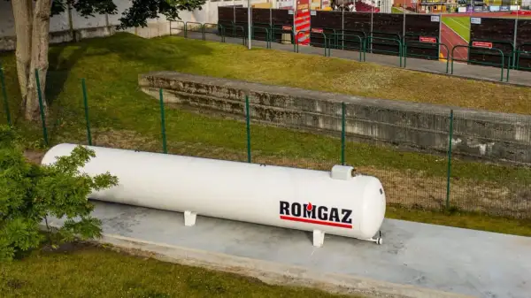 Biały zbiornik gazu ROMGAZ stoi na betonowej podstawie. Widać ogrodzenie i trawę, a w tle trybuny stadionu. Odległość gazociągu od ogrodzenia jest widoczna.
