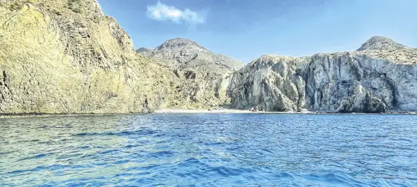 Aguas azules bañan la costa rocosa de una cala en Almería. Montañas escarpadas y una pequeña playa de arena componen el paisaje.
