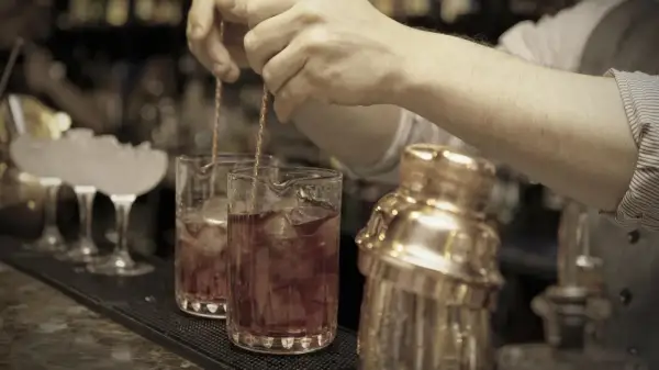 Barkeeper mixt einen roten Bier Cocktail mit Eis in einem Glas. Daneben steht ein goldener Shaker.