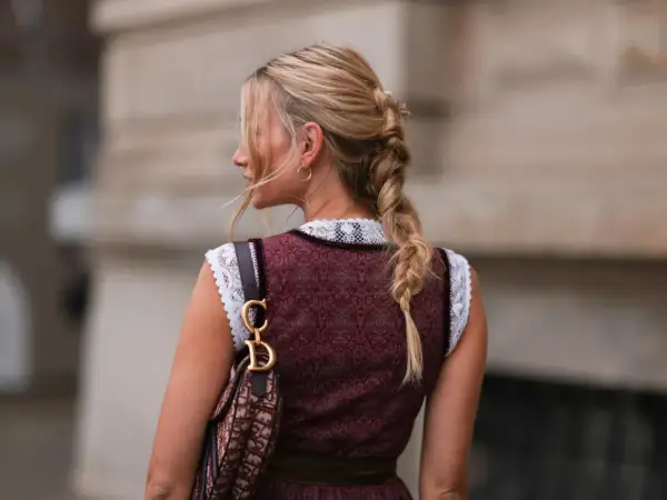 Blonde Zopf-Frisur mit Blumenakzenten, perfekt für Oktoberfest Frisuren. Trägt Dirndl und eine Designer-Tasche.