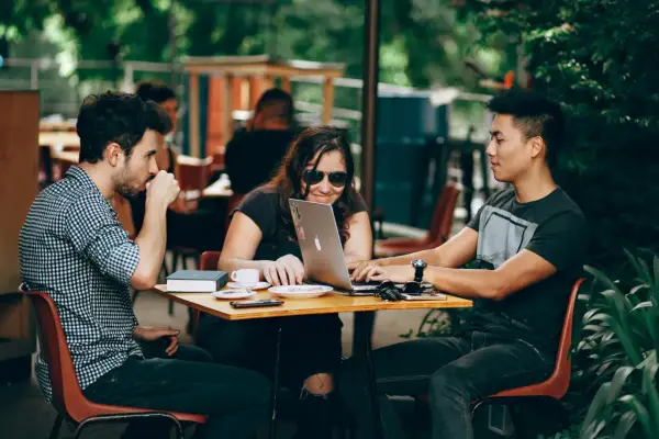 Tres jóvenes colaboran en un portátil en una cafetería al aire libre, un ejemplo de cómo ser nómada digital.