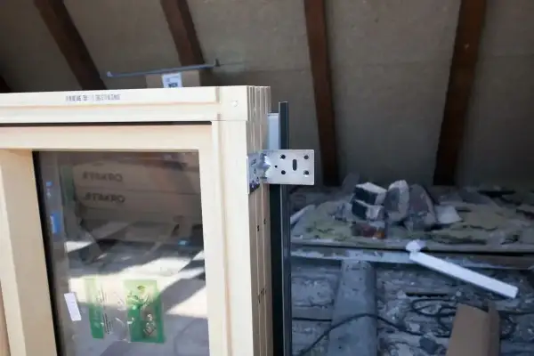 Drewniana rama okna z metalowym wspornikiem, gotowa do montażu. W tle widać materiały budowlane i belki dachu. Okno jest wykonane z materiału podobnego do **pvc**.