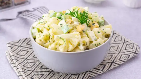 Sałatka z kalafiorem, jajkiem i ogórkiem, udekorowana koperkiem, podana w misce na wzorzystej serwetce.