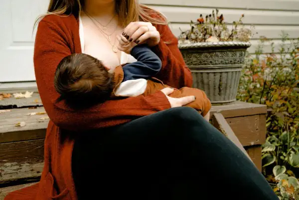 En mamma ammar sitt barn utomhus. Hon bär en röd kofta och mörka byxor. Bilden ger en känsla av lugn och närhet, och kan inspirera till att gå ner i vikt amning på ett hälsosamt sätt.