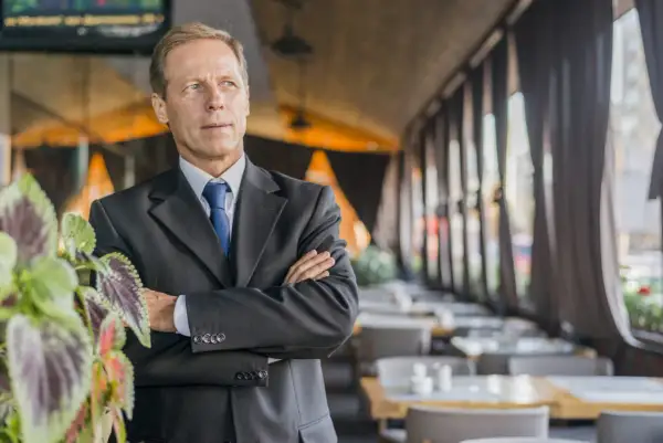 Uomo d'affari con braccia conserte, un chief people officer che guarda lontano, con piante verdi in primo piano e un ristorante sullo sfondo.