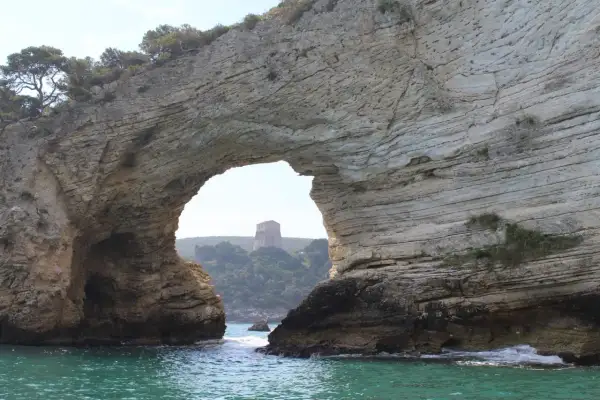 Arco naturale di roccia che incornicia una torre sulla collina, vista dalla baia San Felice Vieste. Acque turchesi e vegetazione mediterranea.