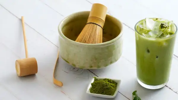 Przygotuj pyszny matcha latte! Na stole leży bambusowa miotełka, łyżeczka, miseczka z zielonym proszkiem matcha i szklanka orzeźwiającego napoju.