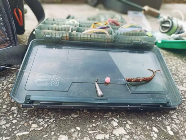 Gotowy jig rig na okonie: przynęta, różowa kulka i ciężarek na żyłce, leżą na otwartym pudełku wędkarskim.