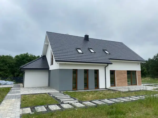 Nowy dom z szarym dachem i białymi ścianami, gotowy do odbioru. Wokół ścieżki z kostki brukowej i trawa.