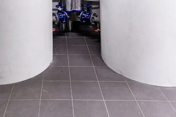 Szare płytki w kotłowni, między białymi zbiornikami. W tle widać instalację hydrauliczną z niebieskimi zaworami.
