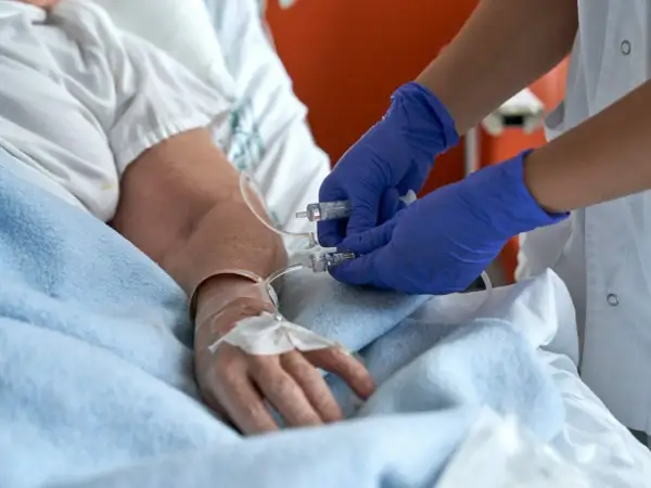 Pielęgniarka w niebieskich rękawiczkach przygotowuje się do usuwania cewnika w domu pacjenta, podłączając strzykawkę do linii infuzyjnej.