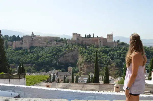 Una mujer contempla la Alhambra, un lugar imprescindible que ver en Granada. El palacio se alza majestuoso sobre un monte verde.