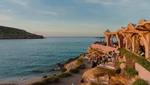 Menschen genießen den Sonnenuntergang am Strand von Ibiza, in der Nähe eines einzigartigen ashram-ähnlichen Gebäudes am Meer.