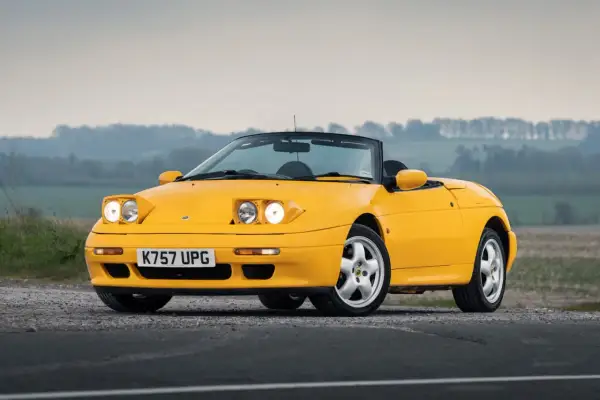 Gul Lotus Elan M100 cabriolet parkerad på grusväg med dimmig bakgrund.