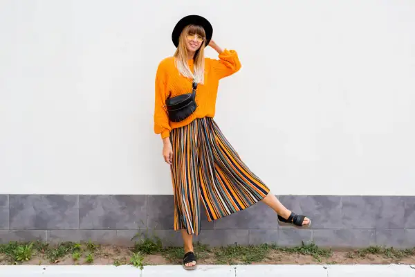 Kobieta w pomarańczowym swetrze i kolorowych kulotach plus size, czarnym kapeluszu i okularach przeciwsłonecznych pozuje na tle białej ściany.