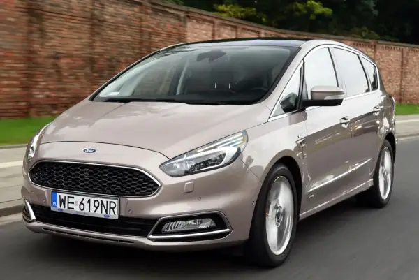 Elegancki Ford S-Max w kolorze piaskowym jedzie po drodze, z ceglanym murem w tle.
