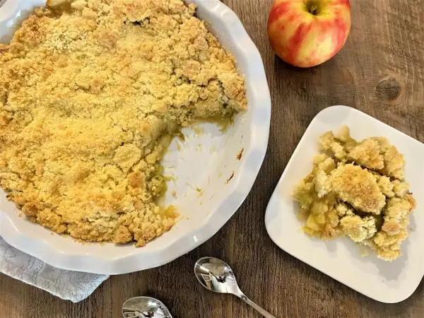 Un délicieux crumble pomme rhubarbe, doré et croustillant, servi dans un plat blanc avec une portion sur une assiette. Une pomme rouge et jaune repose à côté.