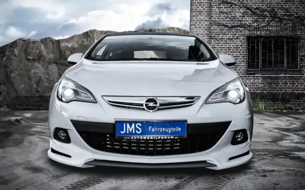 Biały Opel Astra J GTC z tuningiem JMS, z tablicą rejestracyjną "JMS Fahrzeugteile", na tle kamieniołomu i ceglanego budynku.