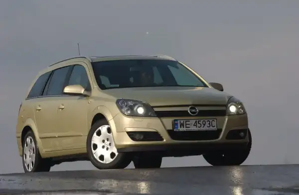 Złoty Opel Astra H kombi z włączonymi światłami. Przód samochodu z wyraźnie widocznymi żarówkami.