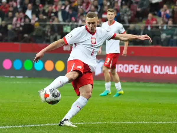 Piłkarz w białej koszulce z orłem i czerwonych spodenkach kopie piłkę na boisku.