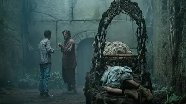 Zwei Männer diskutieren in einer mysteriösen, moosbewachsenen Kammer. Im Vordergrund schläft eine Person in einem rustikalen Bett. Szene aus "Der Greif Staffel 2".