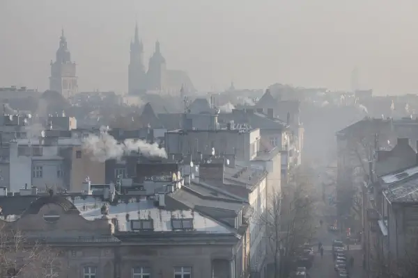 Widok na miasto w smogu z dymiącymi kominami i zarysami wież kościelnych w tle.