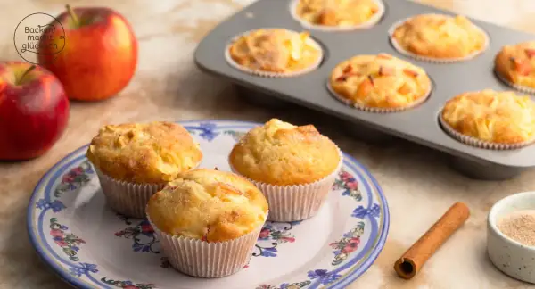Saftige apfelmuffins ohne zucker, frisch gebacken und auf einem Teller mit Blumenmuster arrangiert. Daneben liegen Äpfel und Zimt.