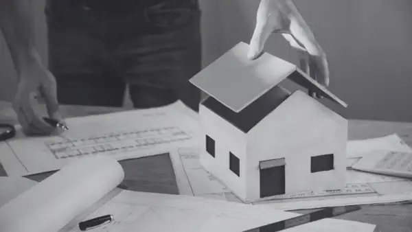 Model domu na papierach z planami. Ręka kładzie dach, symbolizując sprzeciw sąsiada na budowę lub trudności w uzyskaniu pozwolenia.