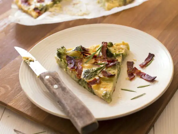 Une part de quiche sans pâte aux brocolis et lardons, garnie de romarin, sur une assiette avec un couteau.