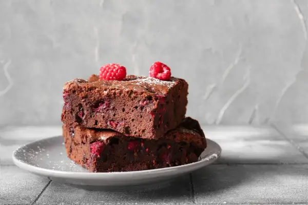 Dwa wilgotne brownie z malinami, posypane cukrem pudrem, na szarym talerzyku. Idealne na deser.
